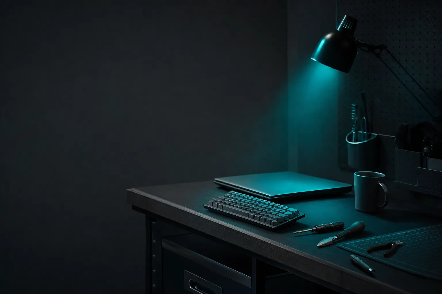 A tidy workbench with a closed laptop, keyboard, tools, and a mug under a teal lamp.
