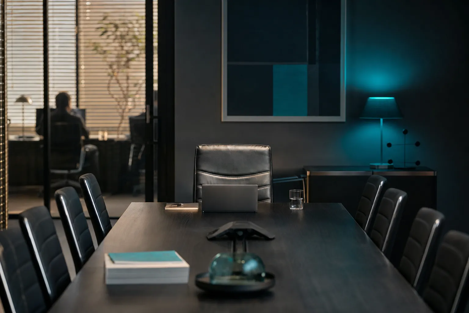 An empty chair at the head of a dark conference table with a laptop and notebook set at the seat.